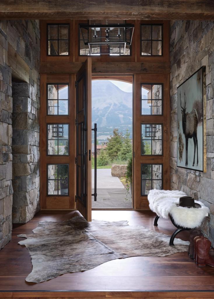 Mountain home entrance in Yellowstone with open door, bench with sheepskin rug and hat on it, painting, views of the mountains in the backdrop
